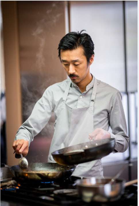 Le chef Atsushi Tanaka cuisine au piano, poêle à la main, dans une cuisine ouverte.