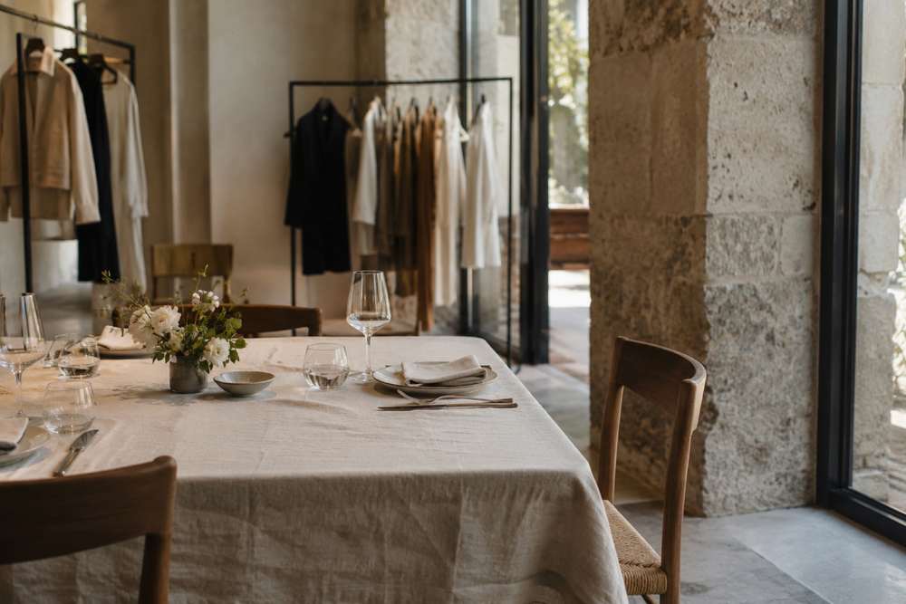 Table de dîner en lin clair et petit bouquet dans une boutique aux portants sobres, éclairée par de grandes fenêtres.